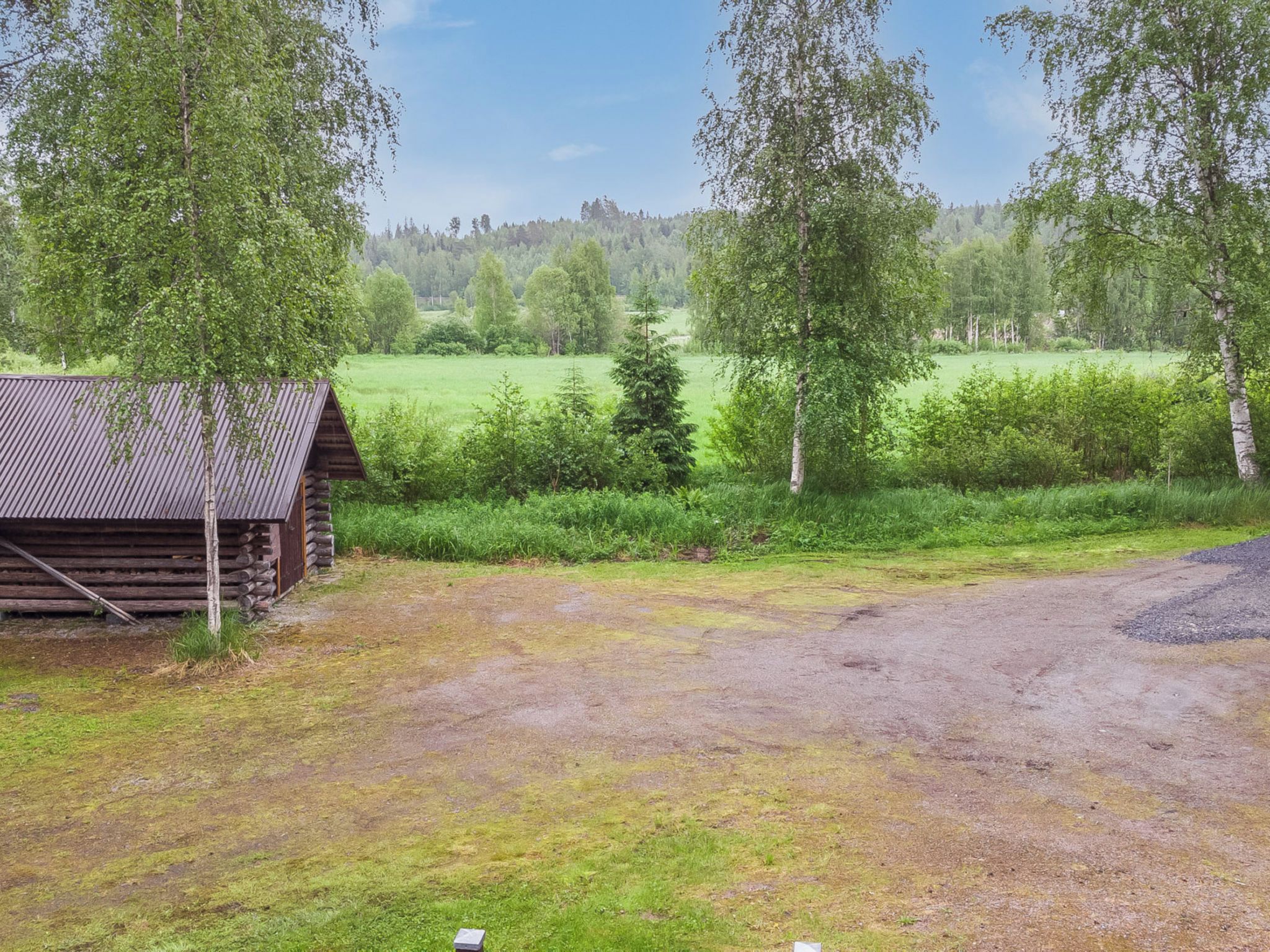 A rustic cabin with a dirt ground in front, surrounded by trees and a grassy field.