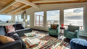 A spacious living room with a large window overlooking the ocean, a cozy seating area, and a kitchen with a view. - Ocean Front Hot Tub, South Beach, Newport, Dog Friendly! (South Beach)