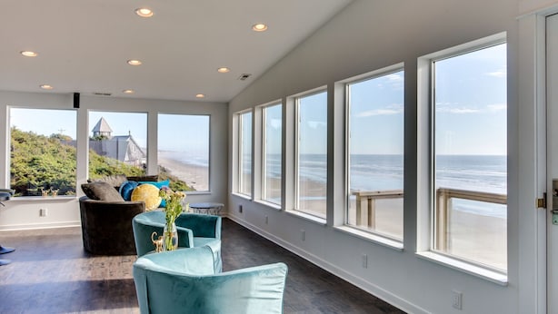 A spacious room with large windows overlooking a beach, a cozy seating area with a table, and a view of a building across the water. - Ocean Front Hot Tub, South Beach, Newport, Dog Friendly! (South Beach)