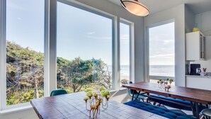 A dining area with a view of the sea and hills, a wooden table, and blue upholstered chairs. - Ocean Front Hot Tub, South Beach, Newport, Dog Friendly! (South Beach)