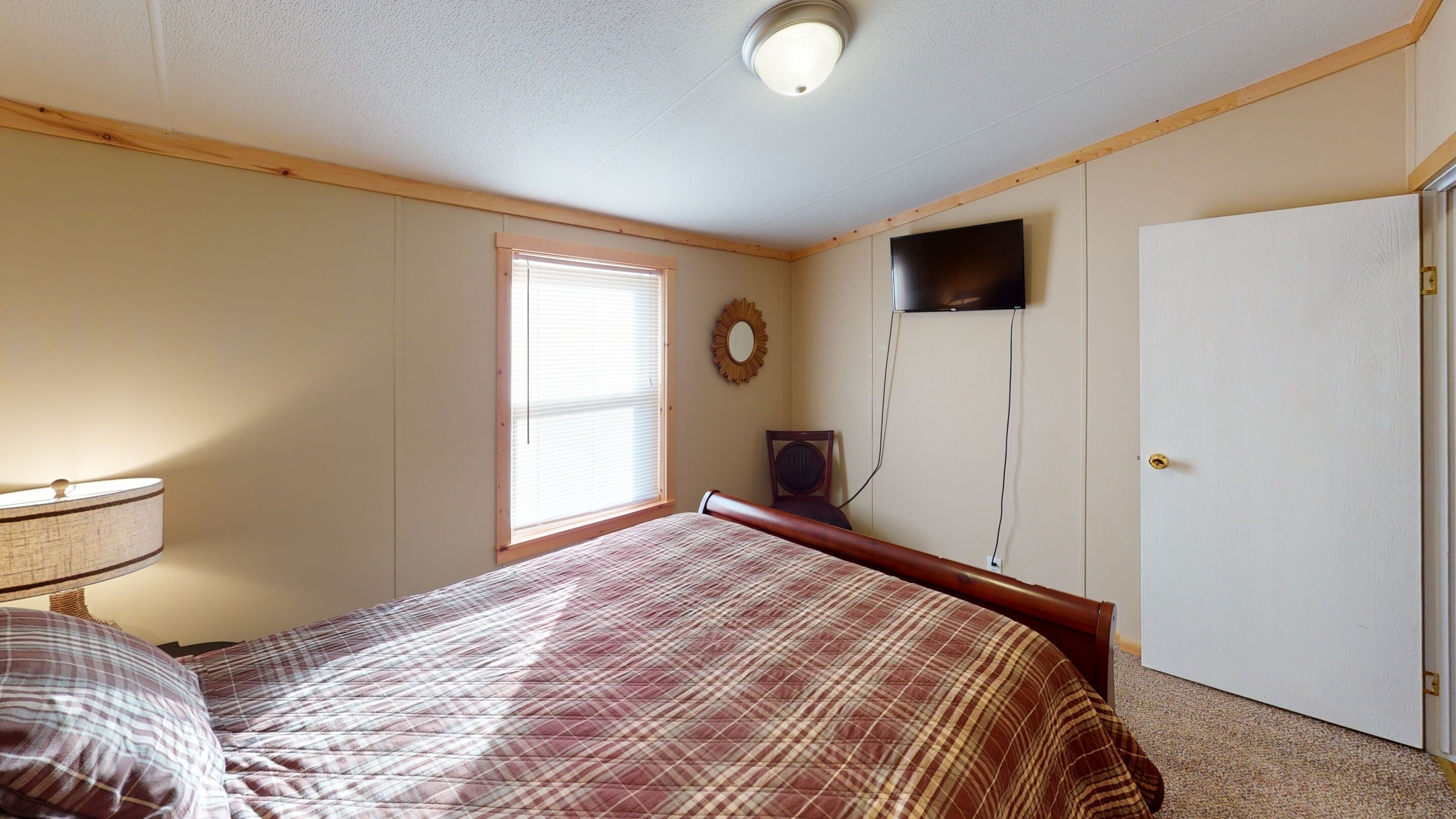 Une chambre à coucher comprenant un lit, une table de chevet, une fenêtre avec des stores, un téléviseur fixé au mur et une porte.