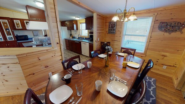 A wooden interior with a dining table set for four, a kitchen area with wooden cabinets, and a window with blinds.