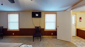 A room with two windows, a ceiling fan, a television mounted on the wall, a chair, and a door leading to another room.