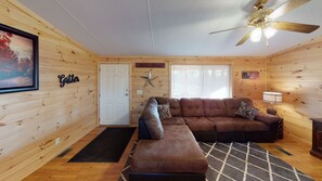 A cozy cabin interior with a brown leather sofa, a wooden door, a ceiling fan, and a framed picture on the wall.