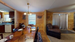 A wooden cabin interior with a dining table, chairs, a sofa, a kitchen area, and a window with blinds.
