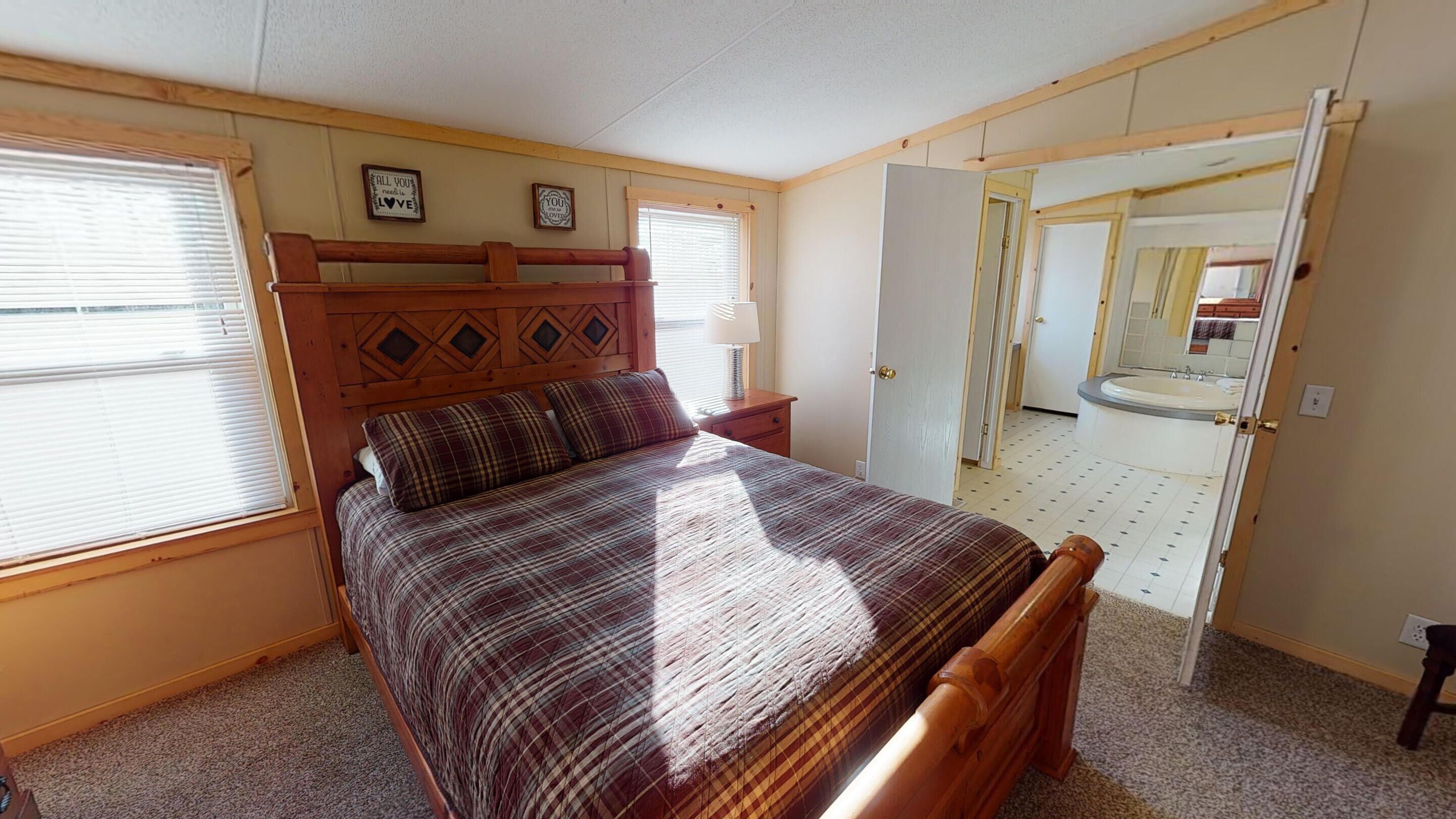 Une chambre avec une tête de lit en bois, une couvre-lit à carreaux et une table de chevet avec une lampe. Il y a deux fenêtres avec des stores, une porte donnant sur une salle de bain et un miroir sur le mur.