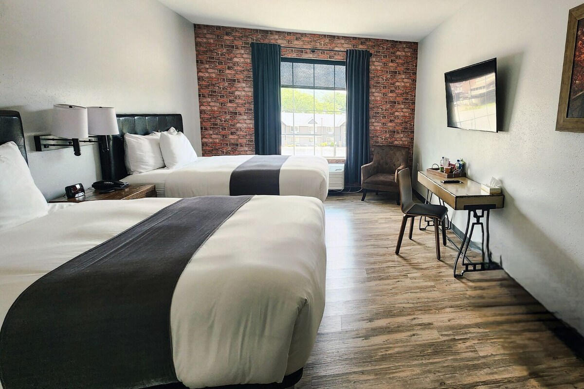 A hotel room with two beds, a desk, and a view of the outside.