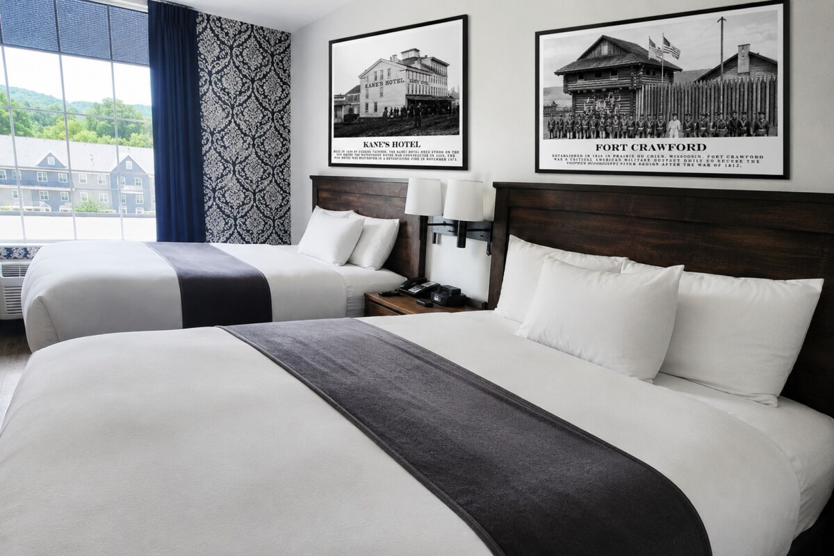A hotel room with two beds, a nightstand with a lamp, and two framed historical photos on the wall.