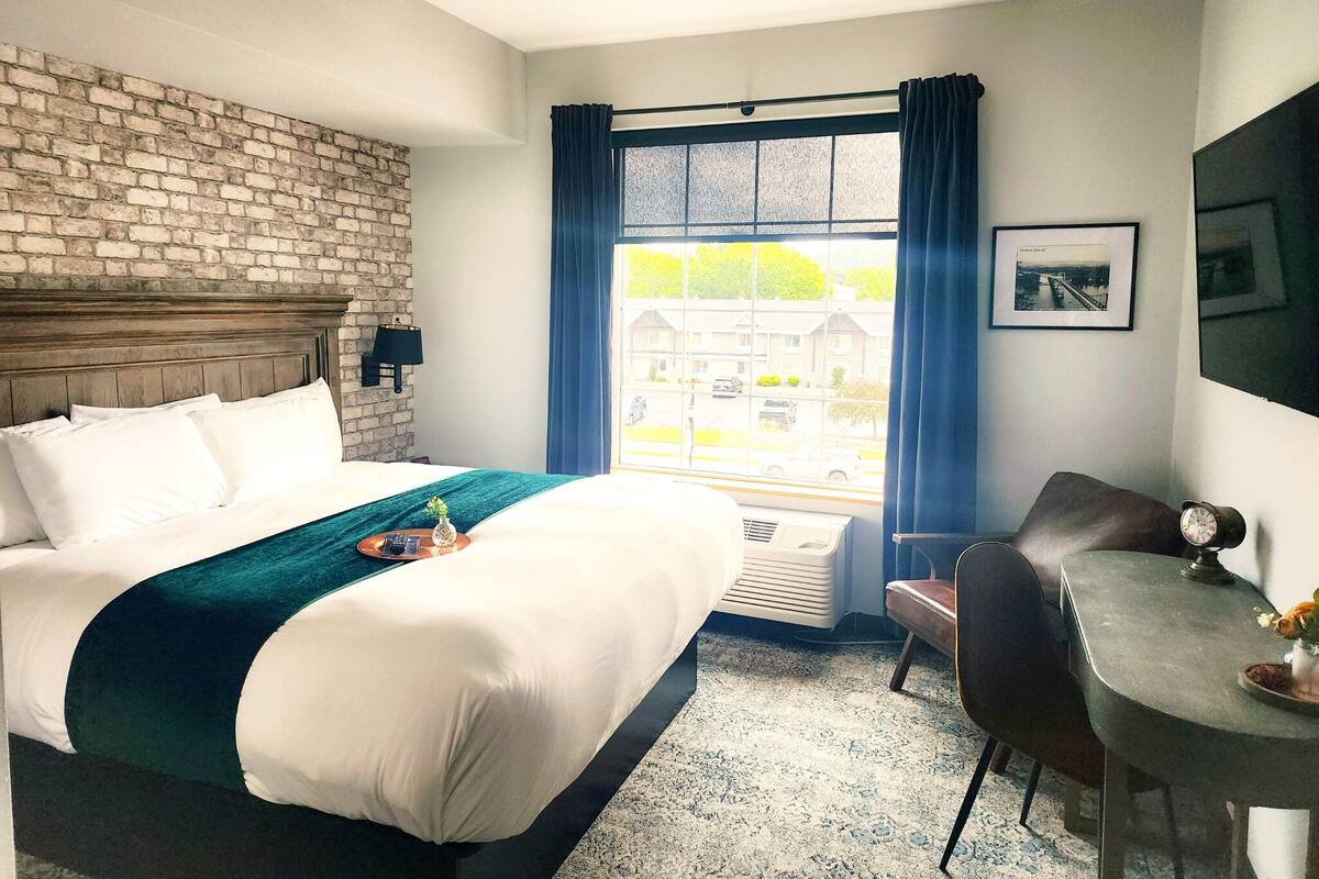 A hotel room with a brick wall, a large bed with white bedding, a desk with a chair, and a window with blue curtains.