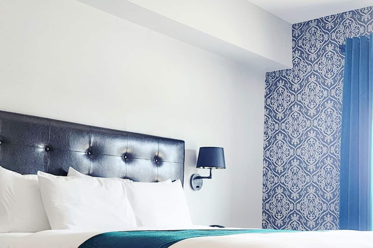 A neatly made bed with white and dark blue bedding, a dark headboard, and a wall-mounted lamp.