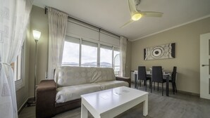 A living room with a sofa, a white coffee table, a dining table with chairs, a ceiling fan, and a view of the outdoors through a large window.