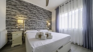 A neatly made bed with white linens and towels, a stone-patterned wall, a white nightstand, and a window with sheer curtains.