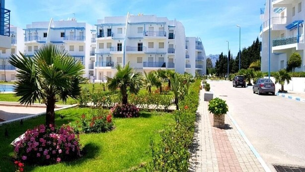 En modern lägenhetsbyggnad med flera balkonger, en välskött trädgård med palmer och blommor samt en klarblå himmel.