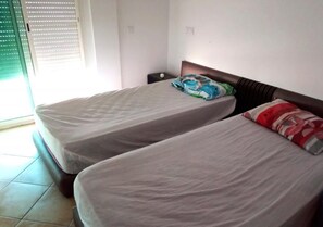 A room with two beds, each with a mattress and a pillow. There is a nightstand with a lamp beside one bed.
