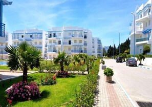 A modern apartment building with multiple balconies, a well-maintained garden with palm trees and flowers, and a clear blue sky. - Residence Riviera Beach 4111 (Cabo Negro)