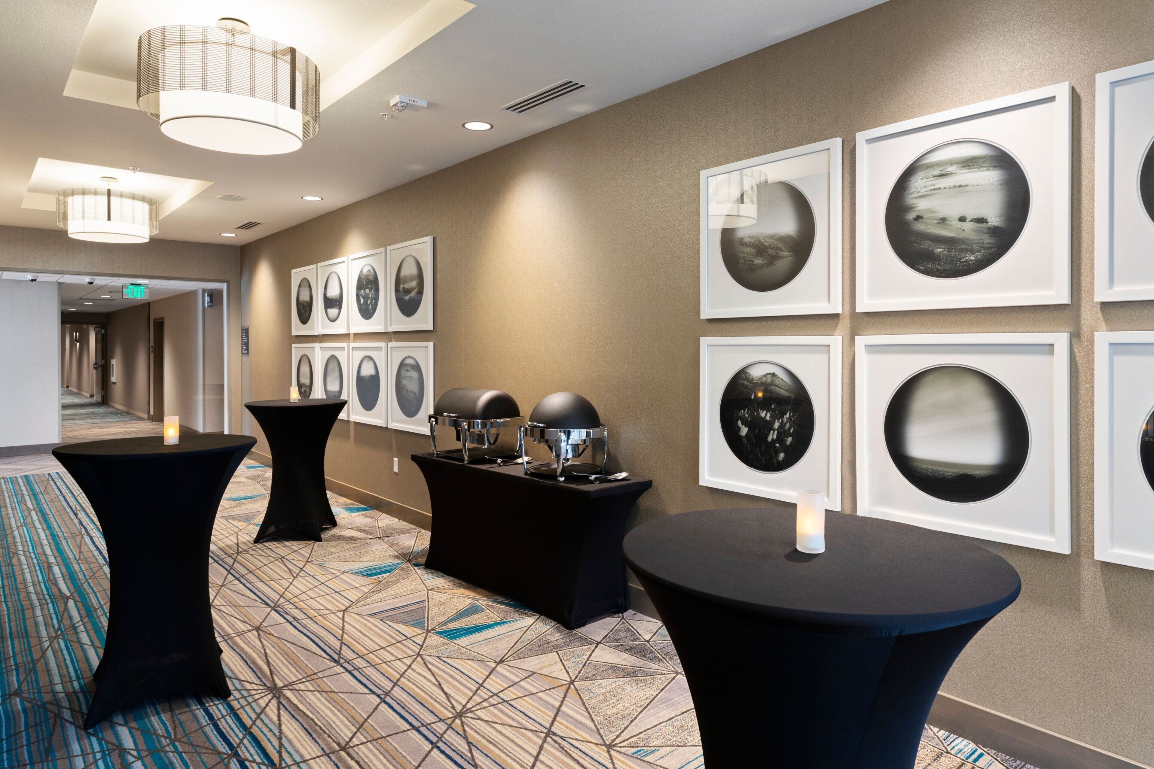 A hotel lobby with a geometric patterned carpet, black round tables with candles, and framed artwork on the walls.
