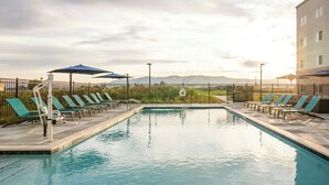 Una piscina rectangular con sillas de descanso y sombrillas, rodeada por una valla, y un edificio al fondo.