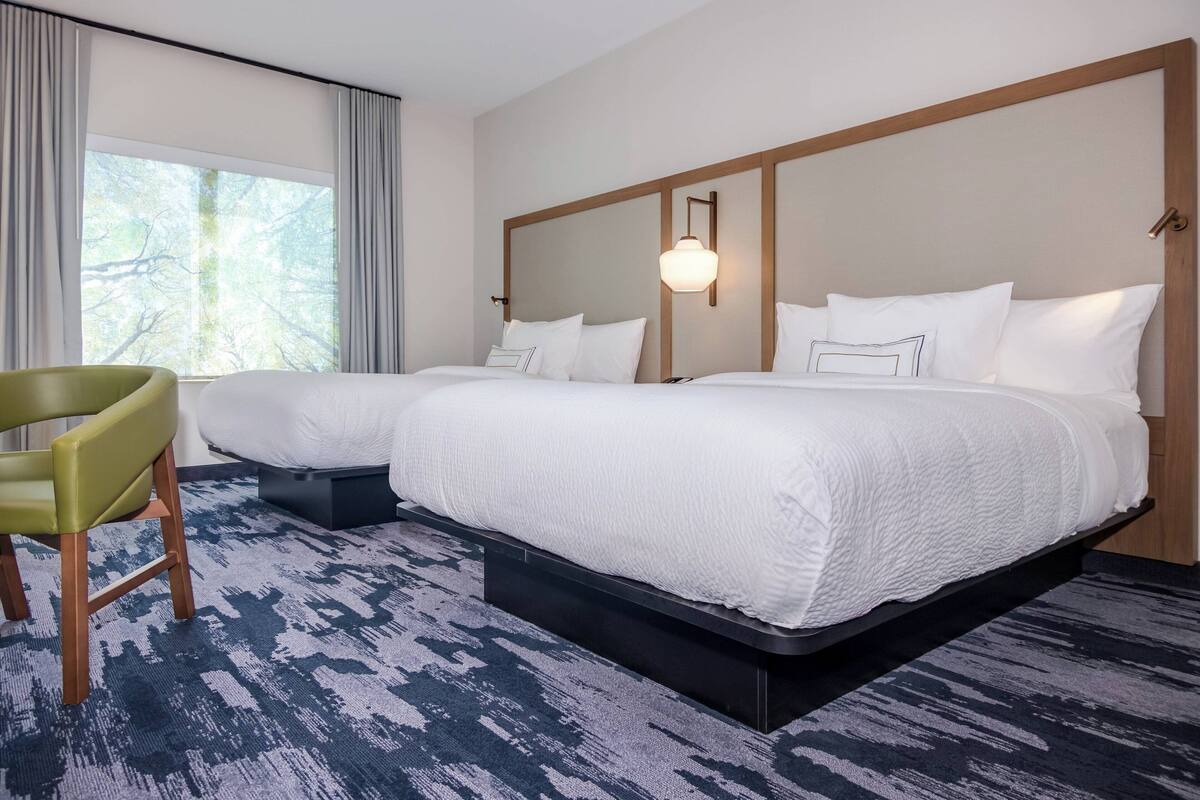 Habitación de hotel con dos camas, una silla verde y un ventanal con cortinas.