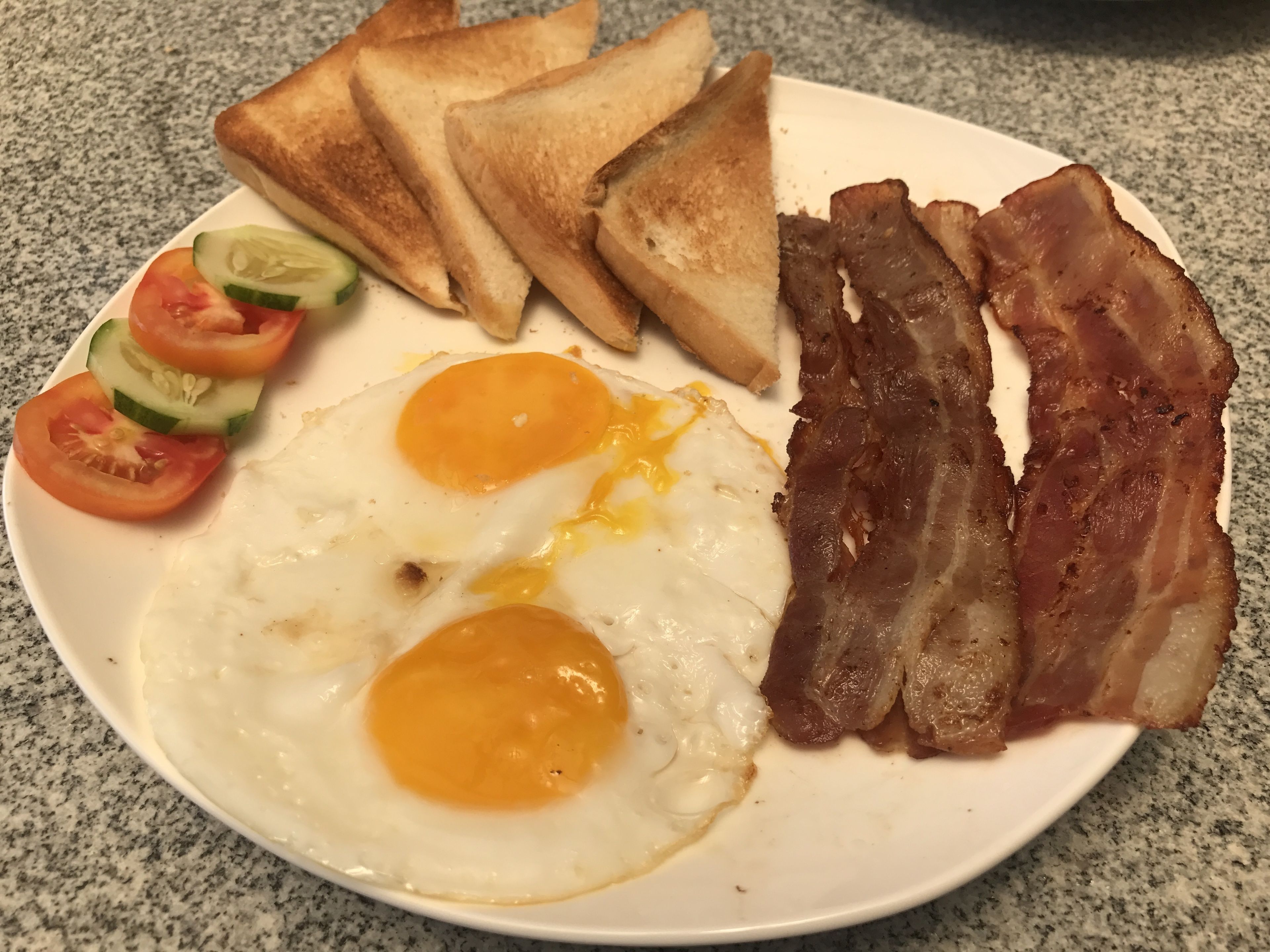 A plate with toast, bacon, eggs, and sliced tomatoes and cucumbers.