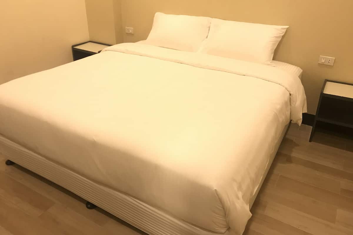 A neatly made bed with white linens in a room with wooden flooring and beige walls.