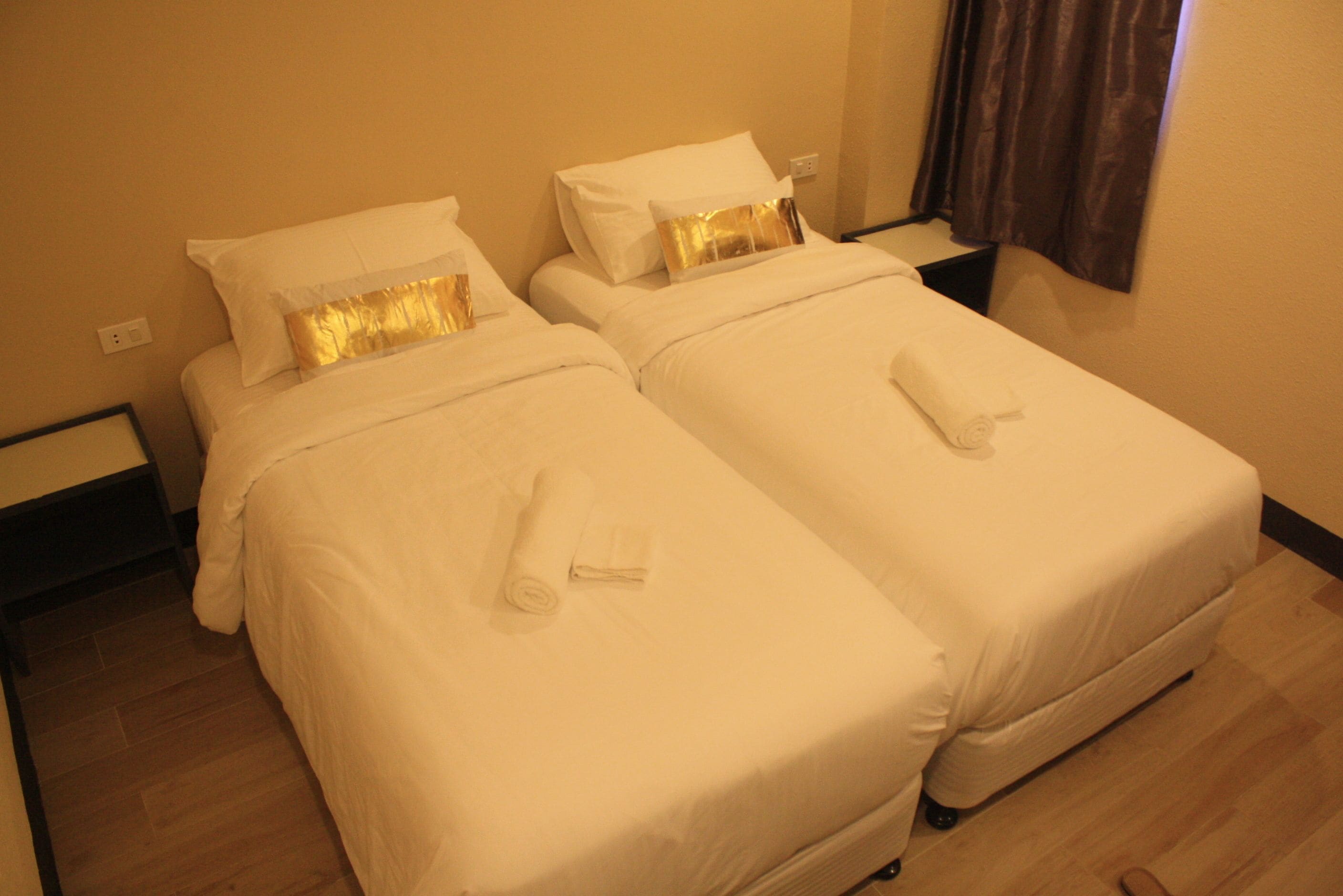 Een hotelkamer met twee eenpersoonsbedden, elk met witte lakens en een gouden sierkussen. Op de bedden liggen opgerolde handdoeken, en de kamer heeft houten vloeren.