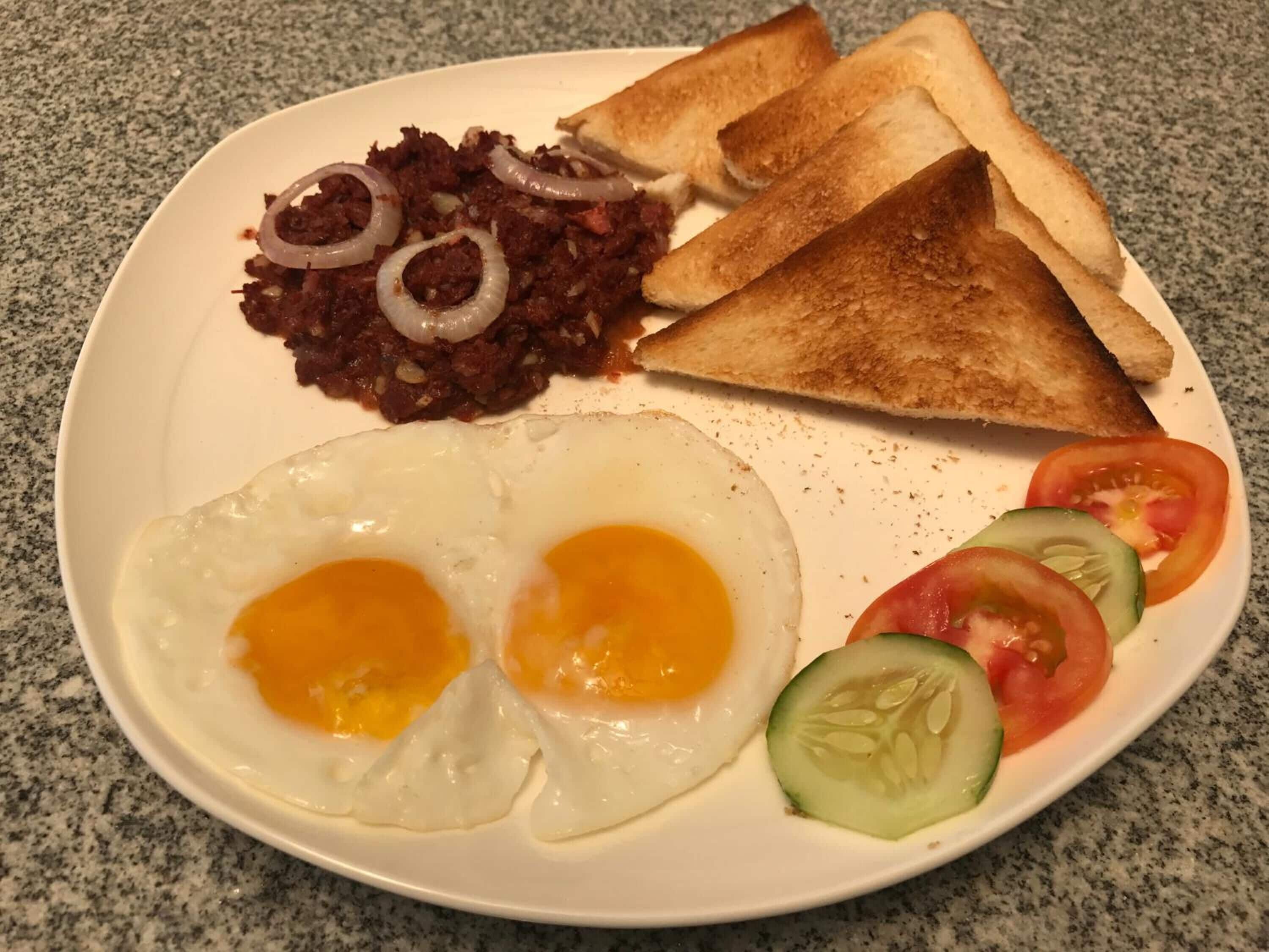 A plate with two fried eggs, toast, a meat dish, and sliced vegetables.