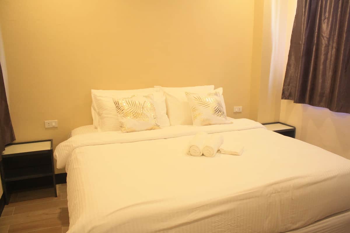 A neatly made bed with white bedding and decorative pillows, a small bedside table, and a power outlet on the wall.