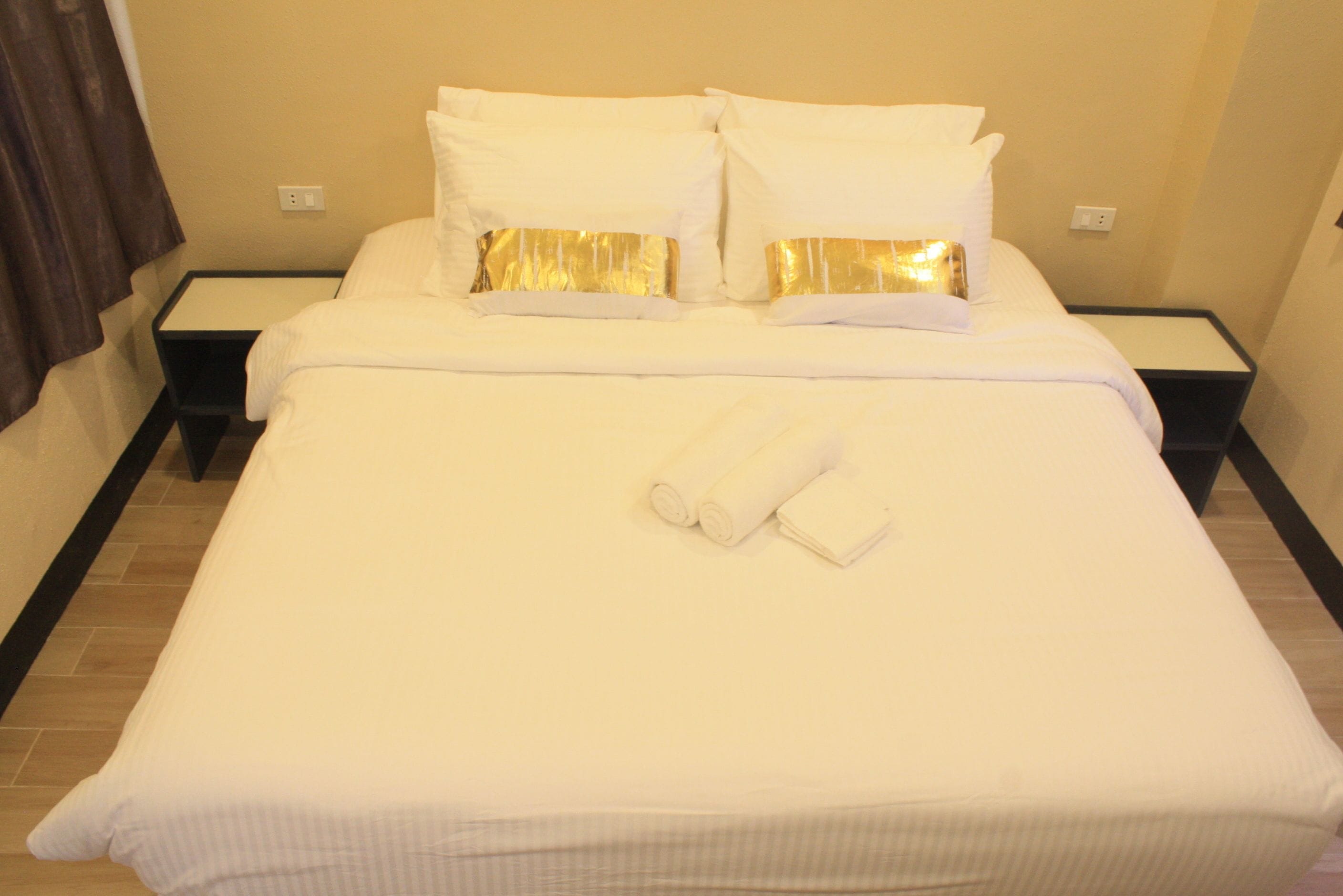 A neatly made bed with white linens and two pillows, a pair of rolled towels, and a black nightstand beside it.