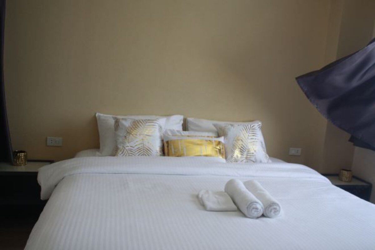 A neatly made bed with white linens, a gold accent pillow, and rolled towels on the bed.