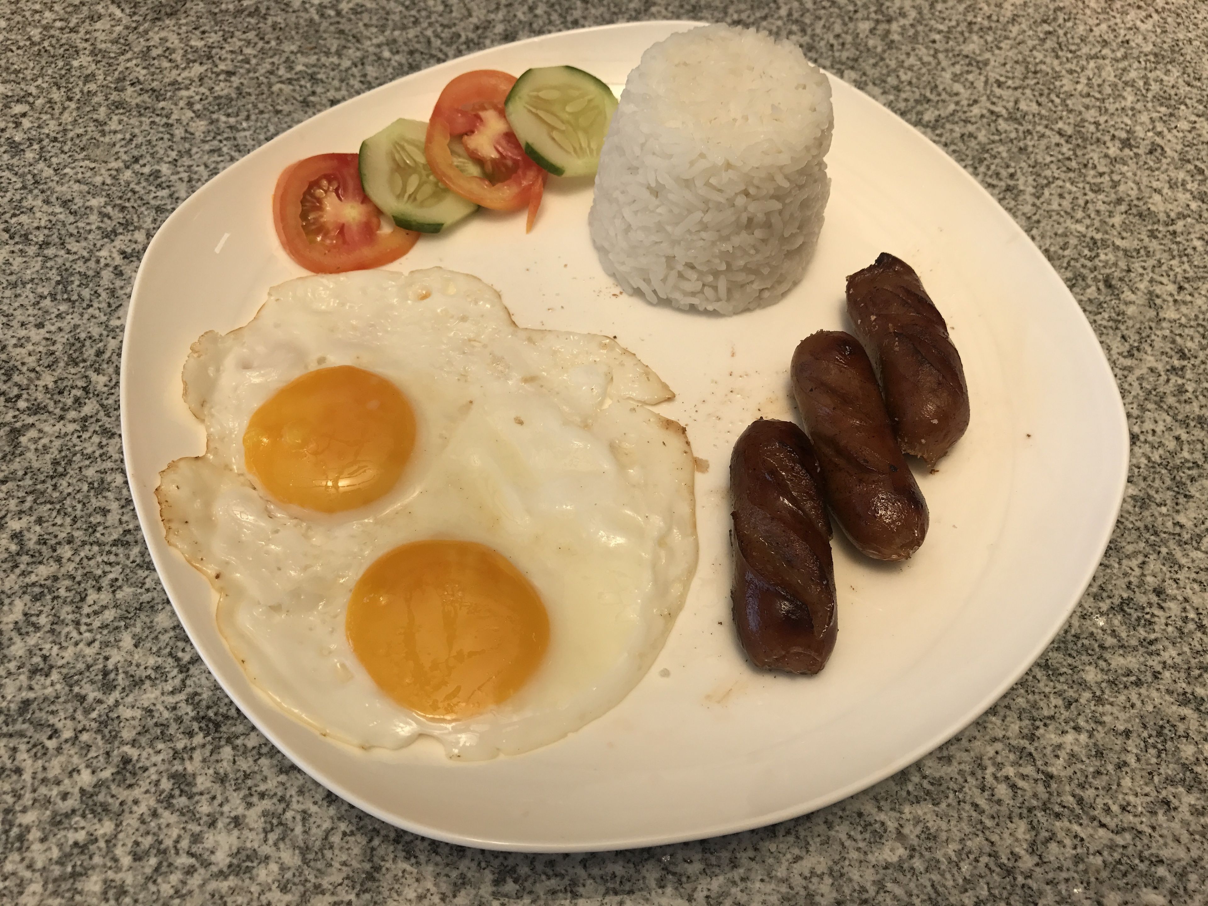 A plate with fried eggs, sausages, rice, and sliced tomatoes and cucumbers.