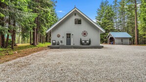 A wooden cabin with a triangular roof, a porch with seating, and a gravel driveway. - Sugar Shack - Peaceful and Private Remodeled Cabin (Leavenworth)