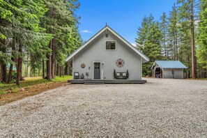 A wooden cabin with a triangular roof, a porch with seating, and a gravel driveway.