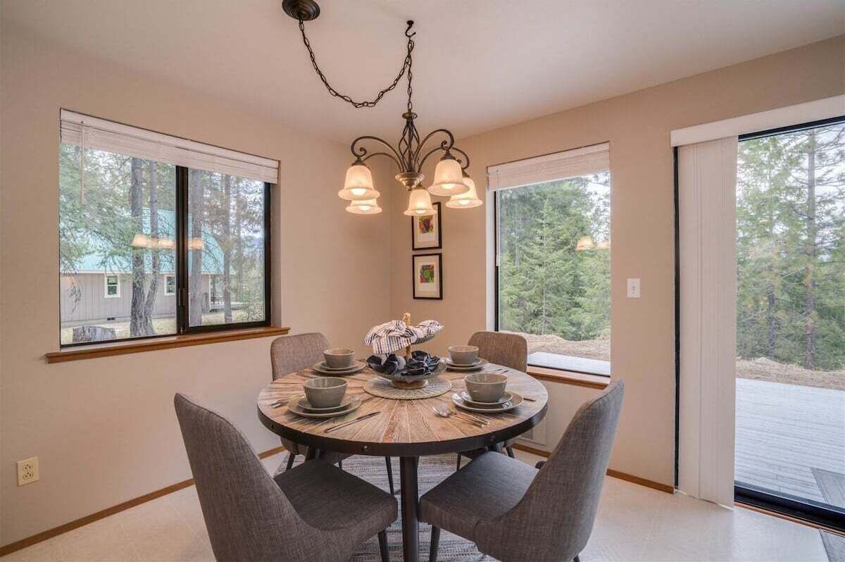 A dining area with a round table, chairs, a chandelier, and large windows with a view of trees.