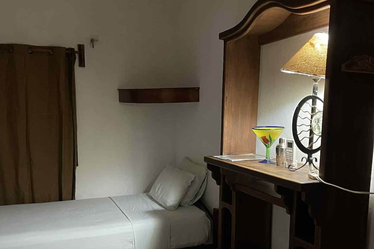Een kleine kamer met een enkel bed, een houten dressoir, een spiegel en een lamp.