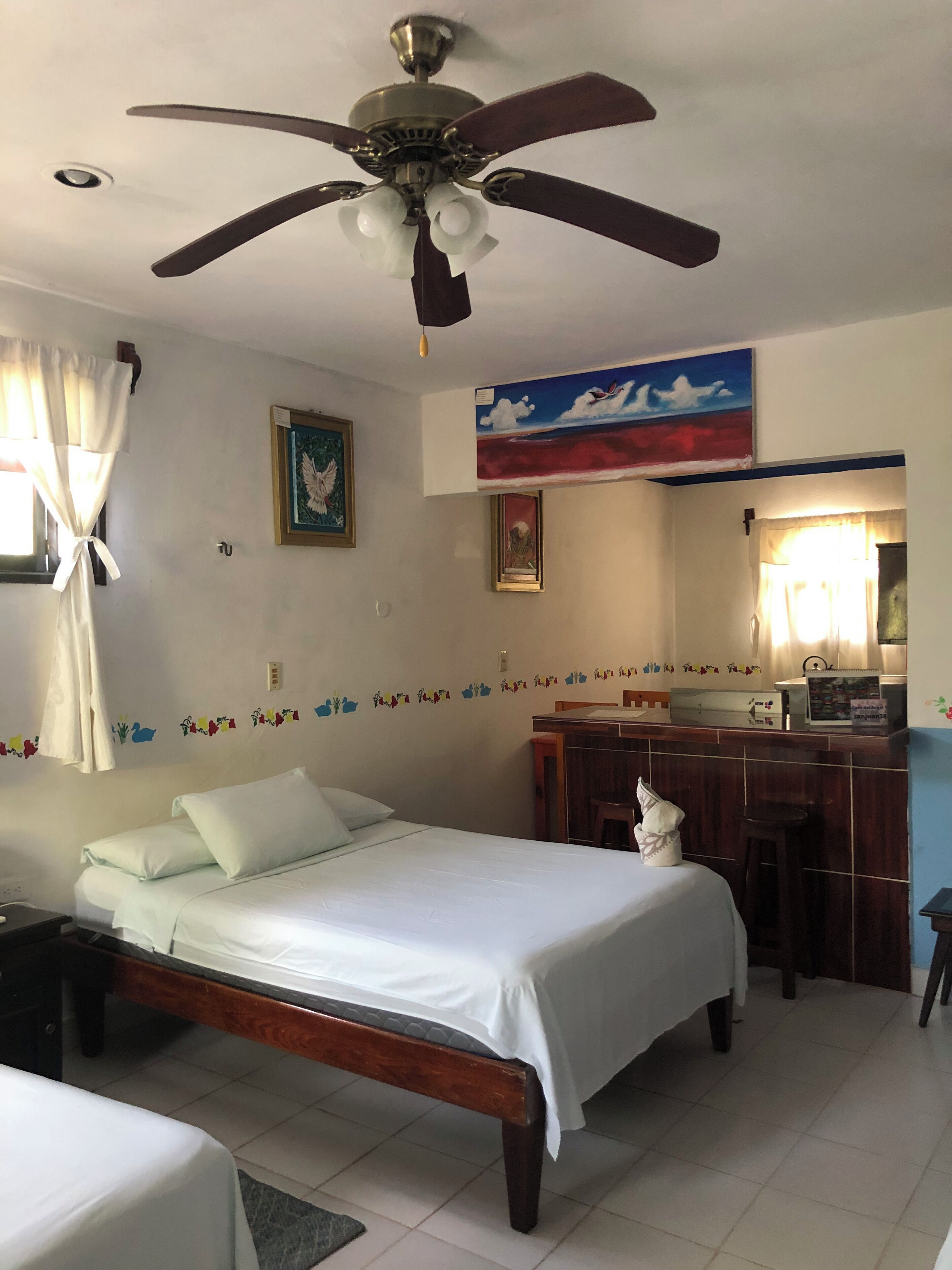 Una camera d'albergo con un letto, una scrivania, una sedia, una ventola a soffitto e una finestra con tende.