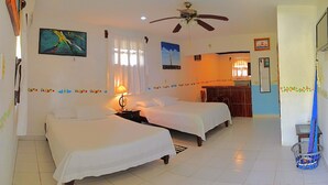 A hotel room with two beds, a ceiling fan, artwork on the walls, and a small kitchenette.