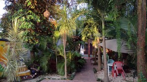 A narrow pathway flanked by lush greenery, including palm trees and various tropical plants, leading to a building with a brick facade.