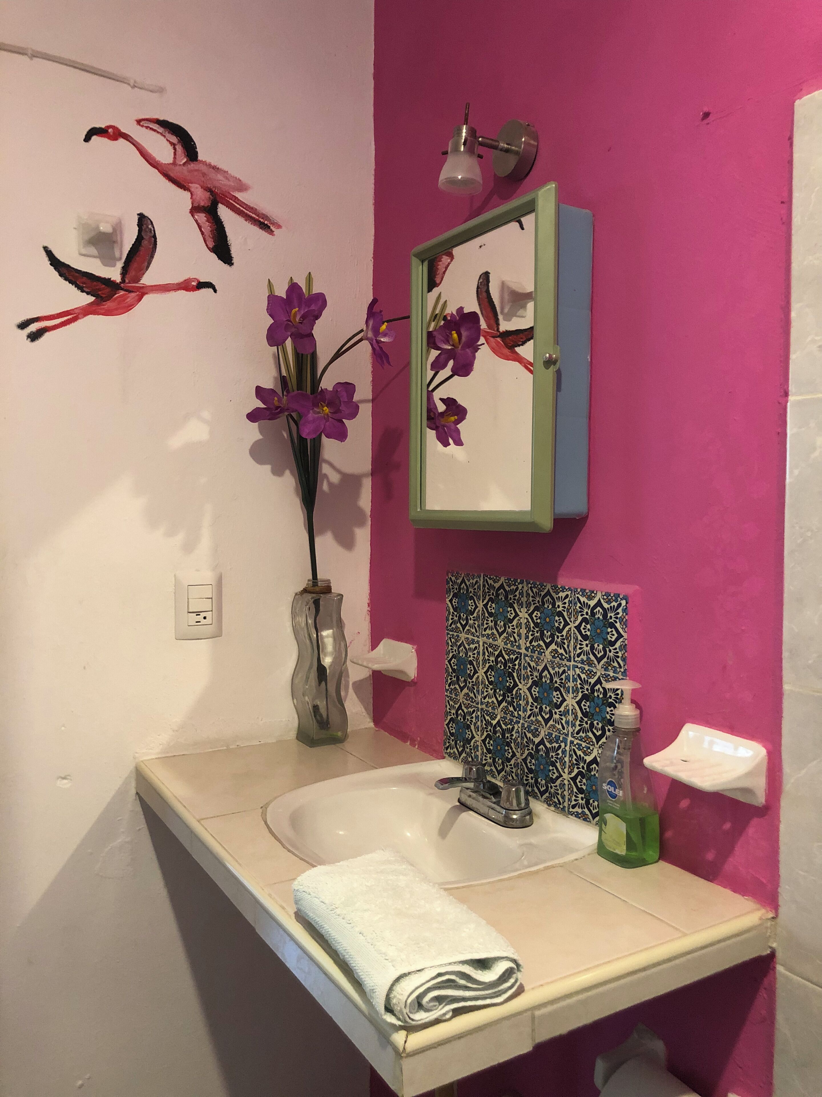 Um banheiro com parede rosa, pia branca, espelho, um vaso com flores roxas e uma toalha no balcão.