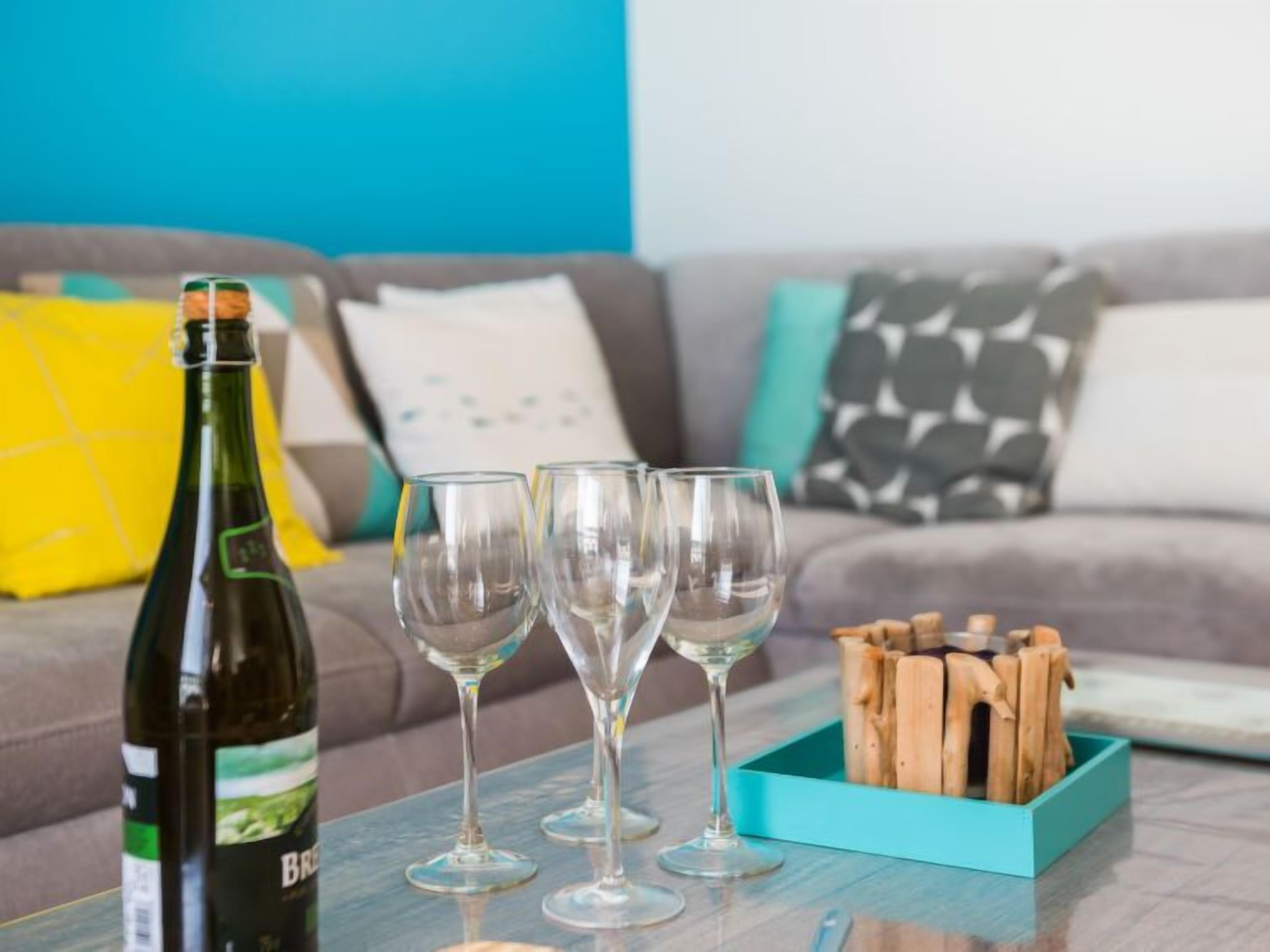 A bottle of wine, three wine glasses, and a box of wooden coasters on a table in a living room with a blue wall and a grey sofa.
