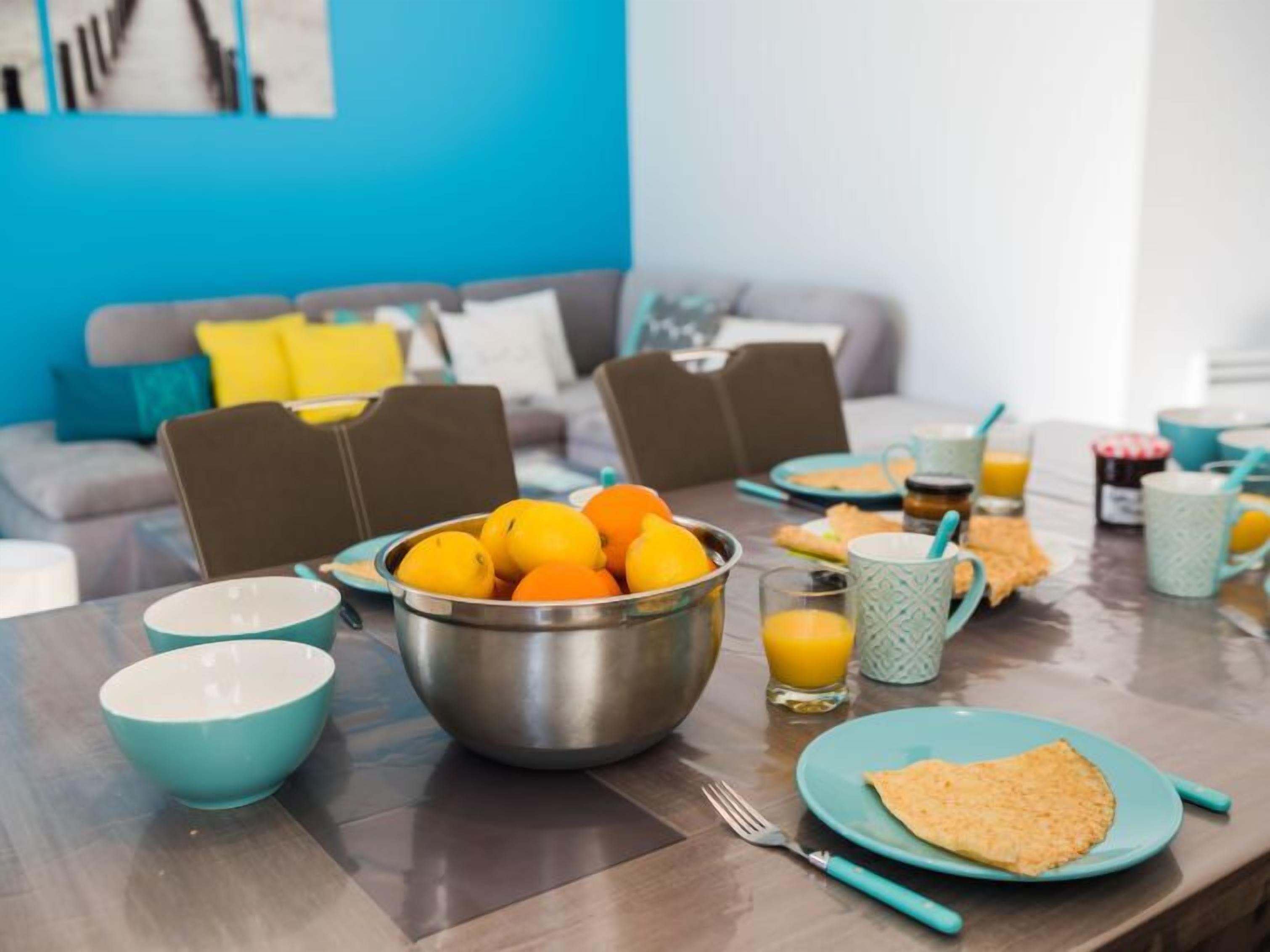 A breakfast setting with a table set for two, featuring a bowl of oranges, a glass of orange juice, and a plate with a folded omelette.