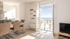 A modern living room with a TV, a dining area with chairs and a table, and a balcony with a view of the sea.
