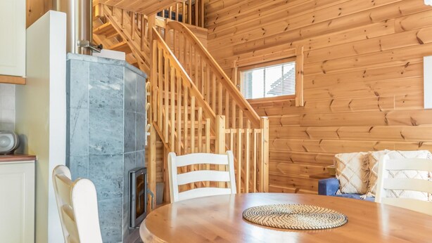 Interior de una cabaña de madera con chimenea de piedra, una mesa redonda de madera y una escalera.