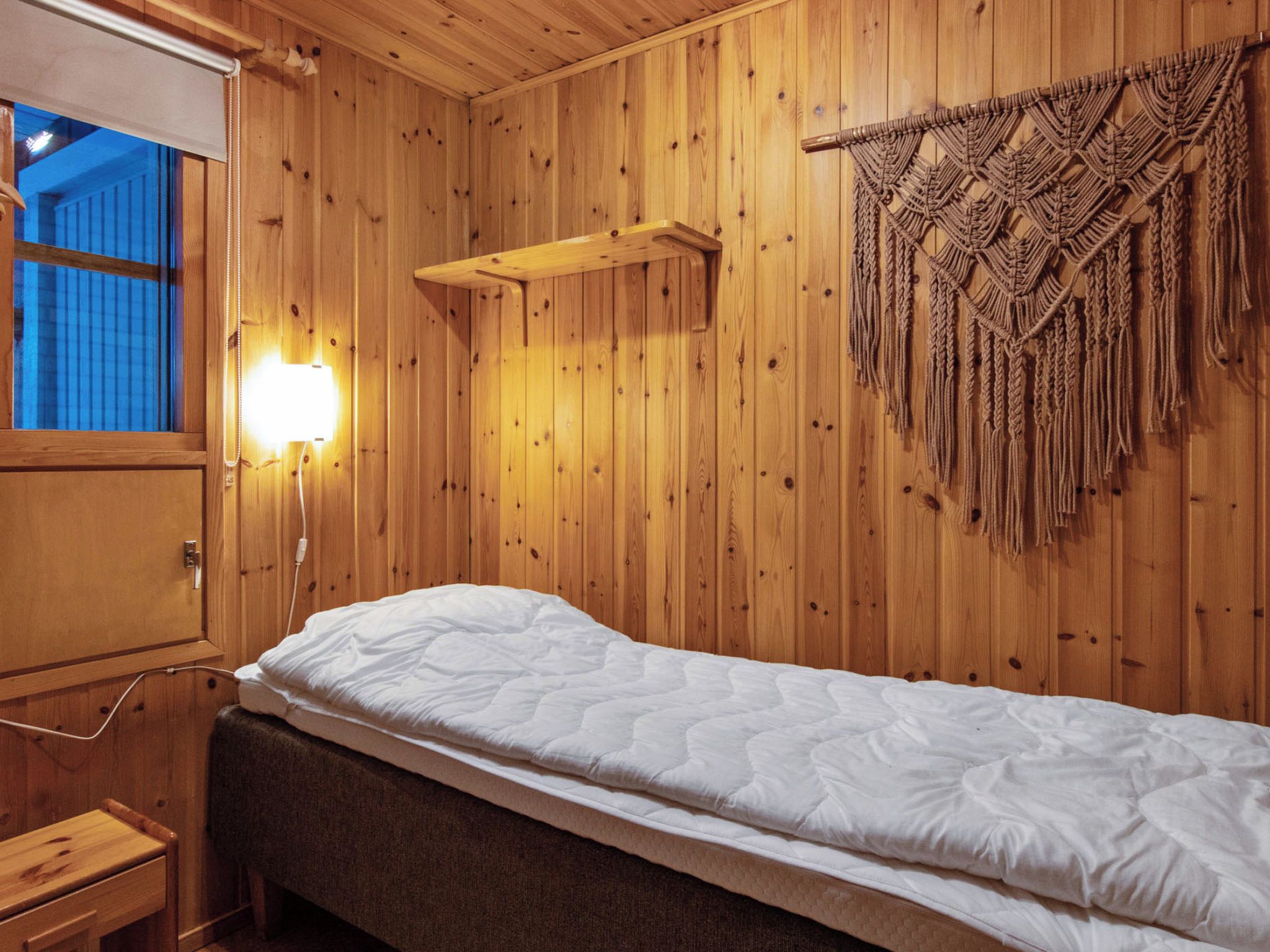 Una habitación de madera con una cama individual, una lámpara de pared y un tapiz de macramé.