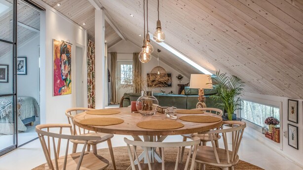 A spacious dining area with a round table and chairs, a ceiling with exposed beams, and a view of a green couch and a plant in the background.