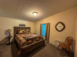 A bedroom with a wooden bed, a bedside table with a lamp, a chair, a window with curtains, and a wall clock. - Spacious Eagle's Landing Cabin near Hocking Hills (Nelsonville)