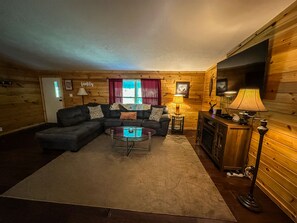 Interior acogedor de una cabaña de madera con un sofá modular, una mesa de centro de vidrio, una chimenea y un televisor montado en la pared.