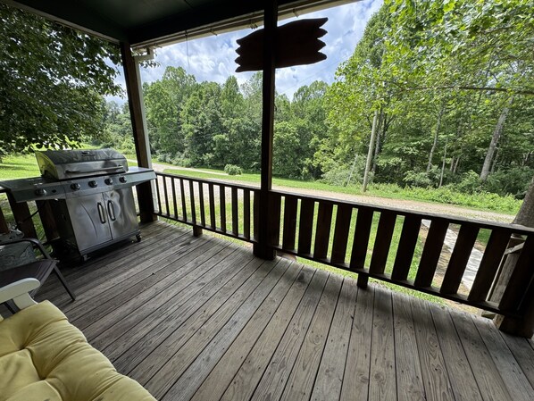 A covered outdoor deck with a barbecue grill, wooden railings, and a view of a wooded area.