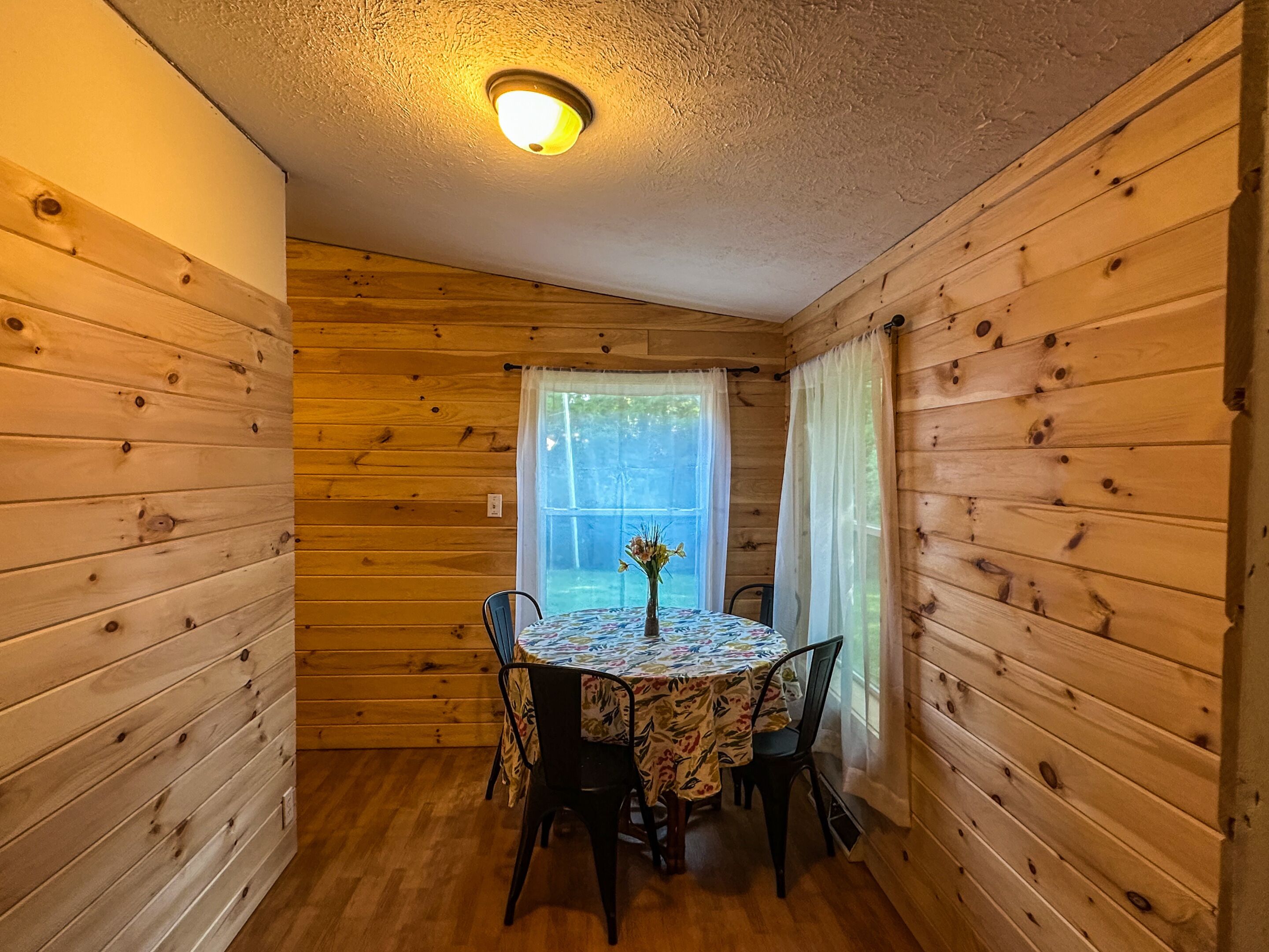 Ein Zimmer mit Holzwänden, einem Tisch, Stühlen, einer Deckenleuchte und einem mit Vorhängen drapierten Fenster.