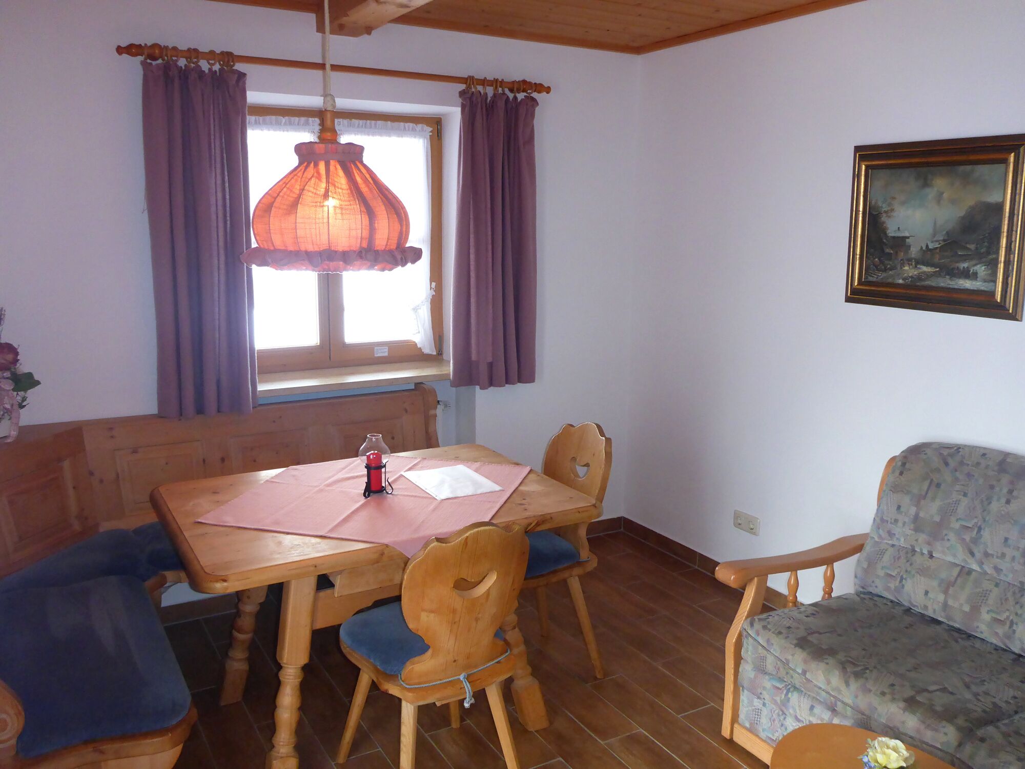 A dining area with a wooden table, chairs, and a bench. There is a window with curtains, a hanging light fixture, and a painting on the wall.