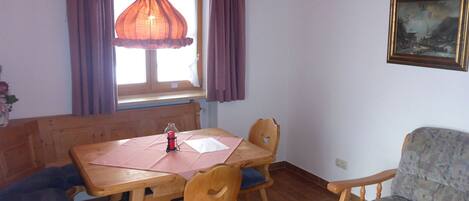 A dining area with a wooden table, chairs, and a bench. There is a window with curtains, a hanging light fixture, and a painting on the wall.